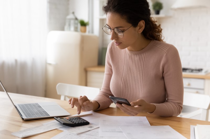 Pesquise os preços e a localização do imóvel | Foto de uma mulher usando o notebook, a calculadora, e o celular para fazer contas | Como comprar uma casa | Blog Alea