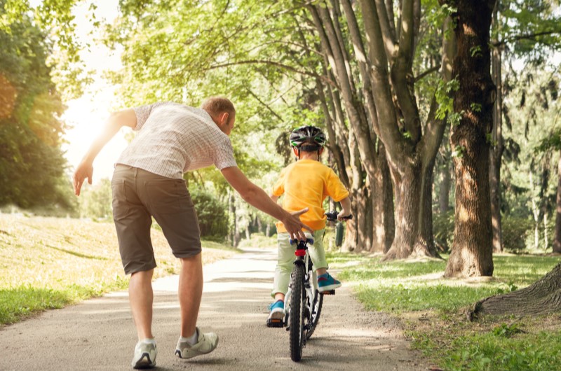 Conheça as opções de lazer da cidade | Foto de um pai ajudando o filho a andar de bicicleta em um parque | Estilo de vida | Blog Alea