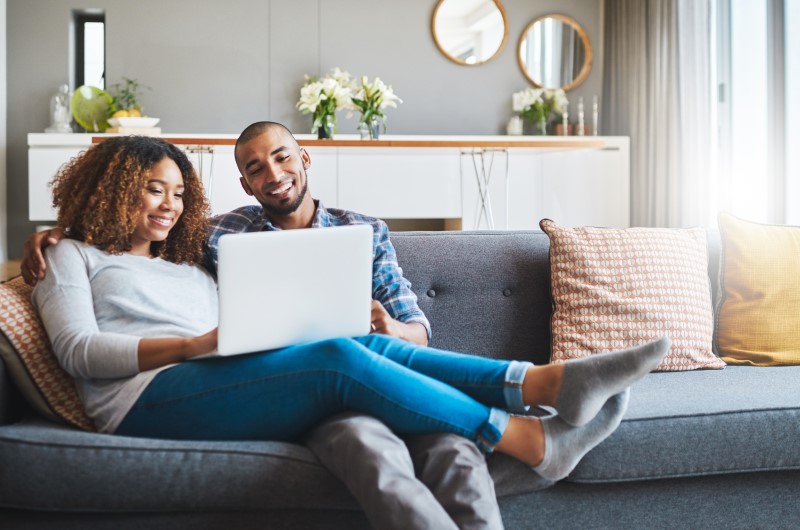 Como é morar em Três Lagoas? | Foto de um casal sorridente sentado no sofá enquanto olham para o notebook | Estilo de Vida | Blog Alea