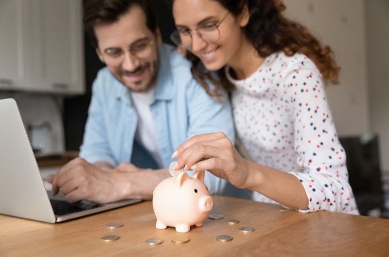 Foto de um casal sorridente em frente ao notebook juntando dinheiro em um cofrinho | O que é melhor amortizar | Como comprar uma casa | Blog Alea