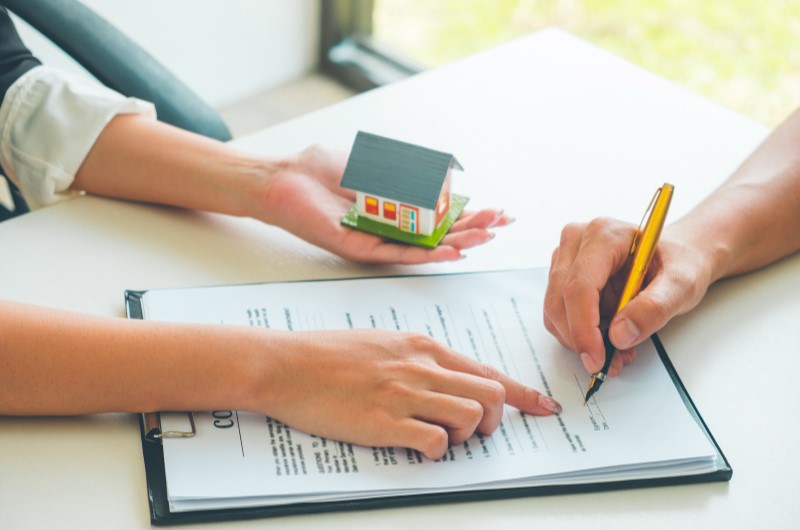 Foto de uma pessoa assinando um contrato e uma pessoa segurando uma casinha e fornecendo informações | Quando é possível fazer a amortização de financiamento | Como comprar uma casa | Blog Alea