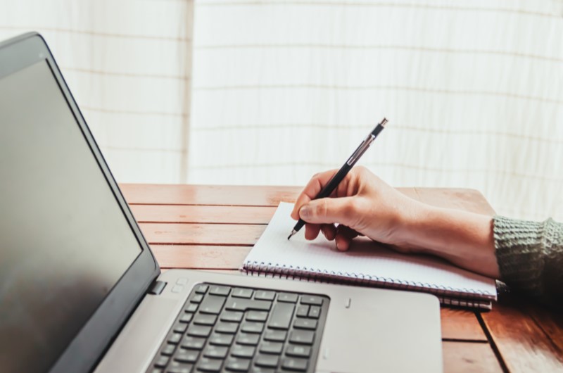 Tabela Price: como funciona? | Foto de uma pessoa escrevendo em uma agenda em frente ao notebook | Como comprar uma casa | Blog Alea