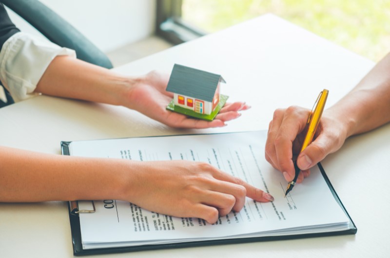 O que é a escritura de imóvel? | Foto de uma pessoa assinando um contrato, enquanto outra pessoa aponta o local no documento | Como comprar uma casa | Blog Alea