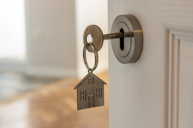 O que é o Minha Casa, Minha Vida? | Foto de uma porta entreaberta, com uma chave e chaveiro de casinha na fechadura | Tudo sobre o Minha Casa Minha Vida | Blog Alea