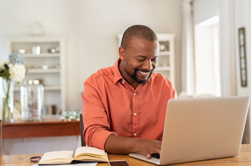 Quais são as vantagens de comprar um imóvel? | Foto de um homem sorrindo e usando o notebook | Como comprar uma casa | Blog Alea